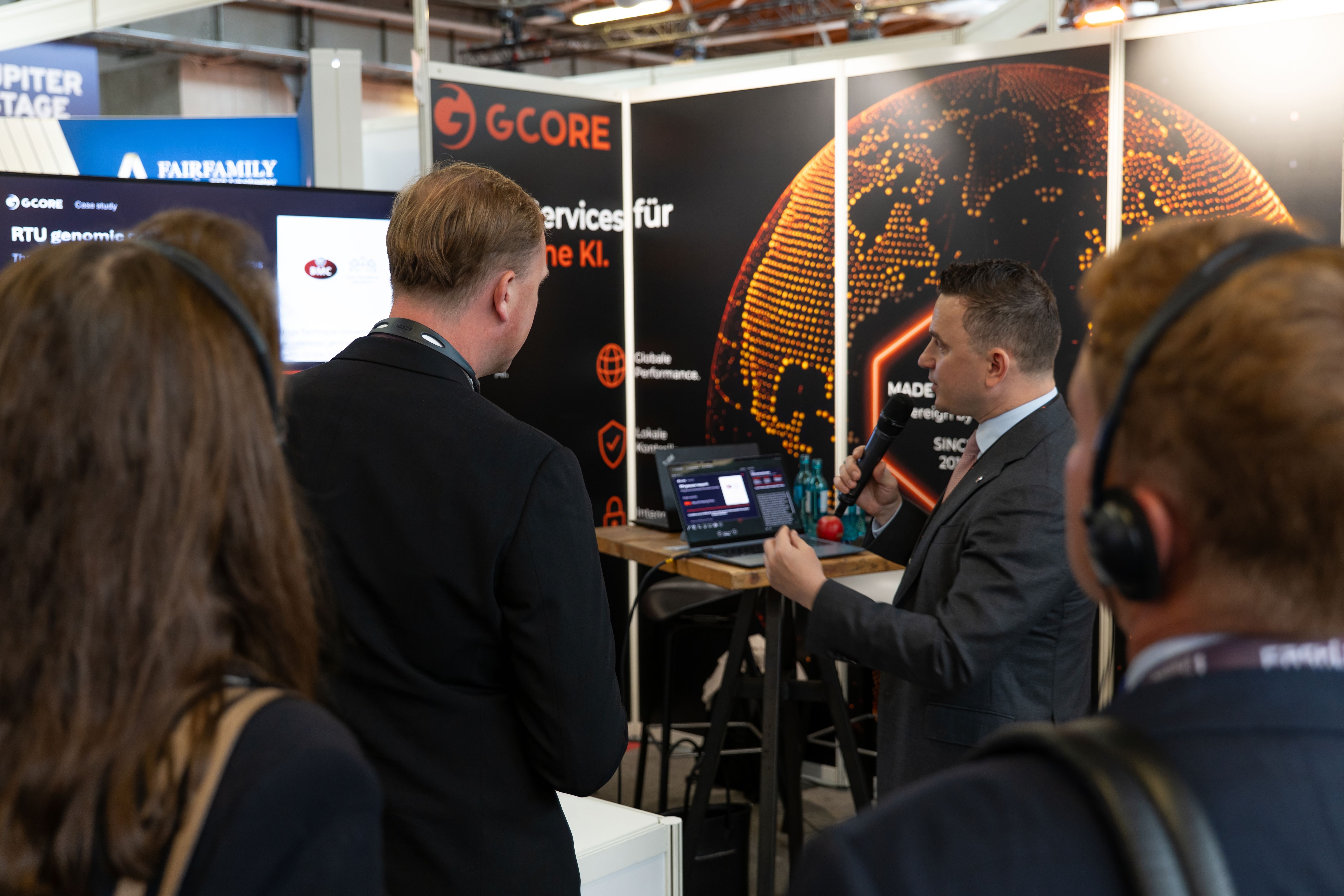 A presenter speaks to an audience at a GCORE booth, showcasing technology on a laptop.