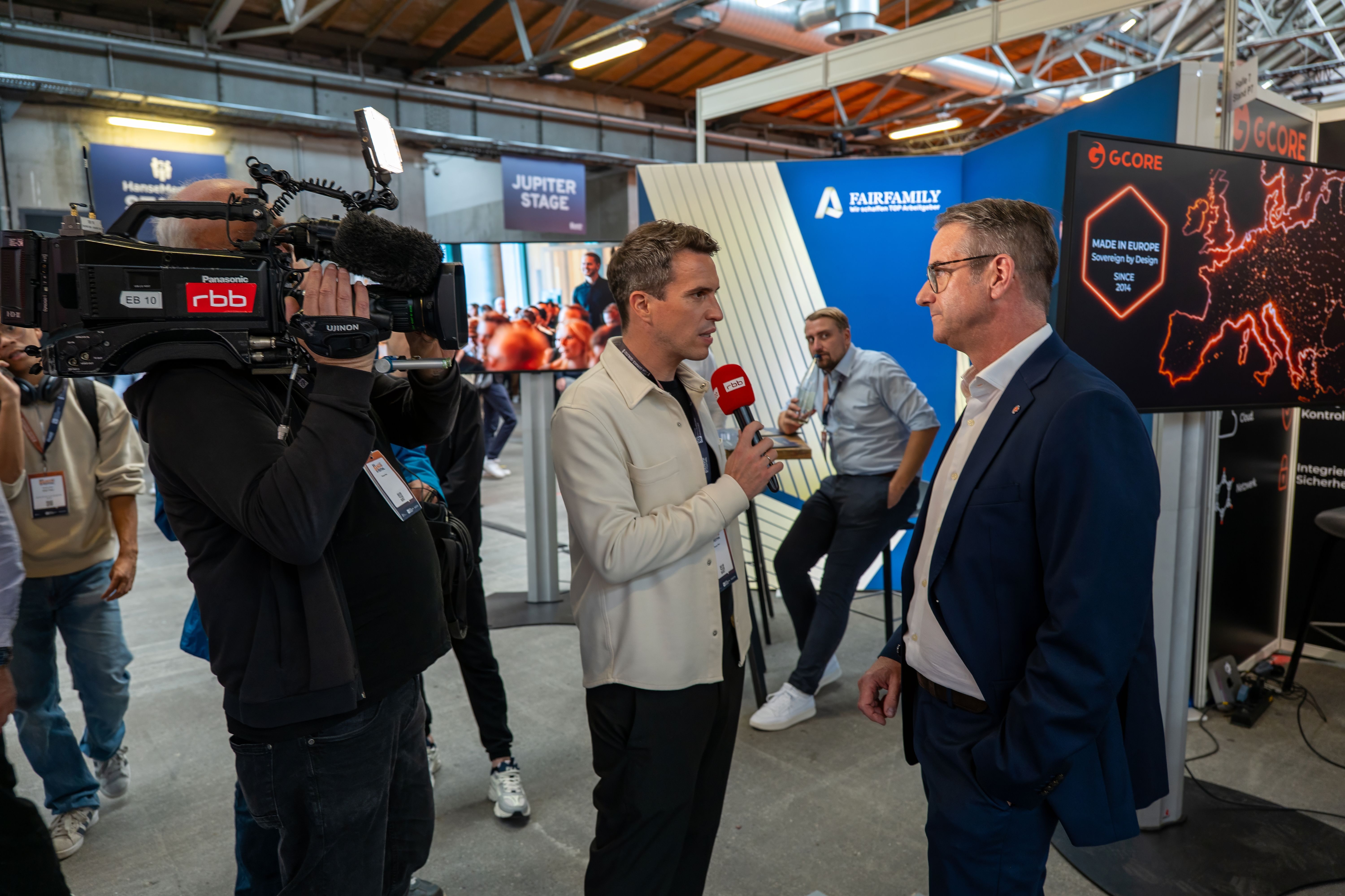 A reporter interviews a man in a suit as a cameraman films at an indoor event.