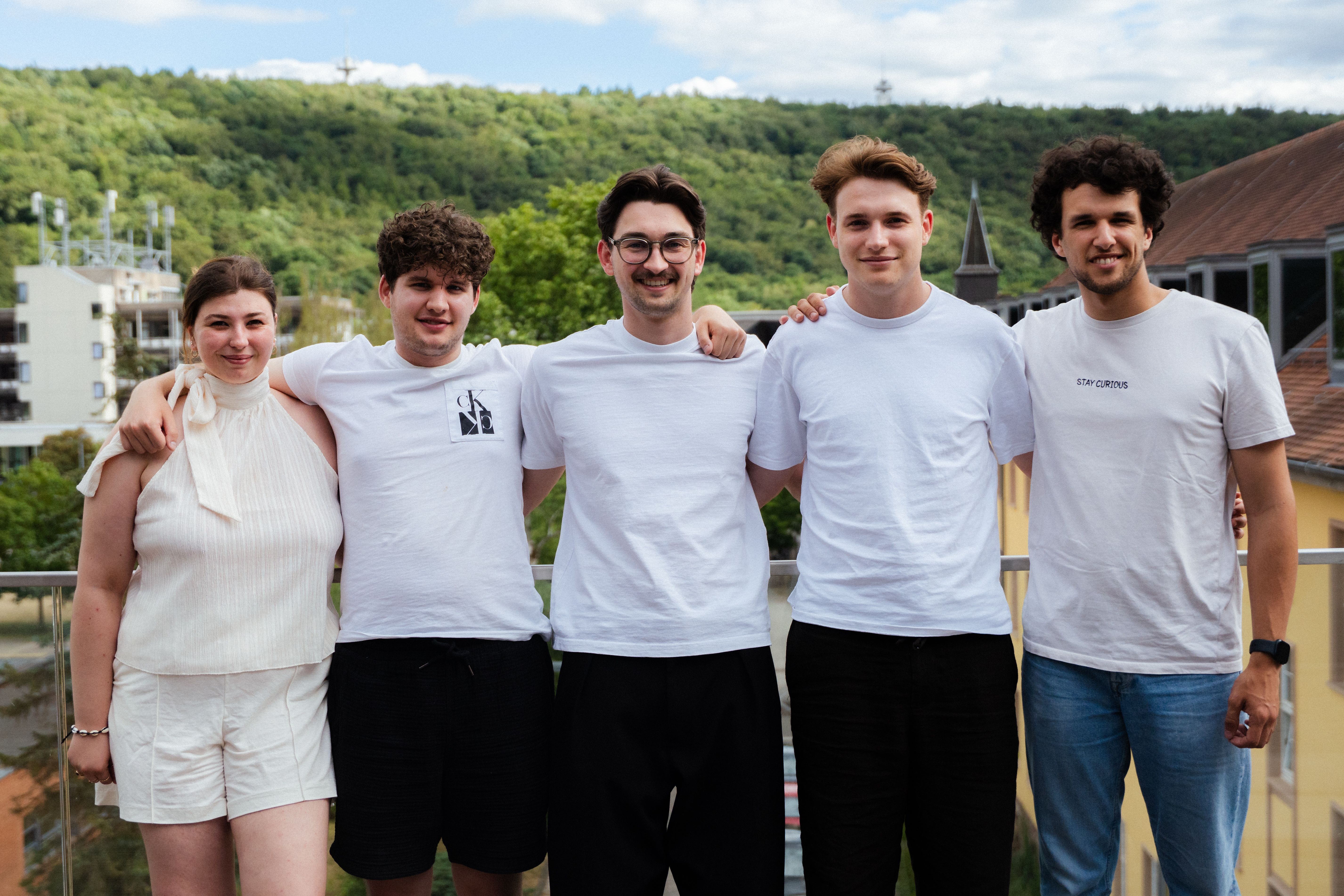 Five smiling young adults standing together outdoors with a green hillside and buildings behind them.