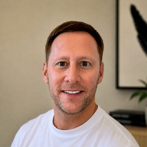 A man with short brown hair and blue eyes smiles warmly, wearing a simple white t-shirt against a light-toned background.