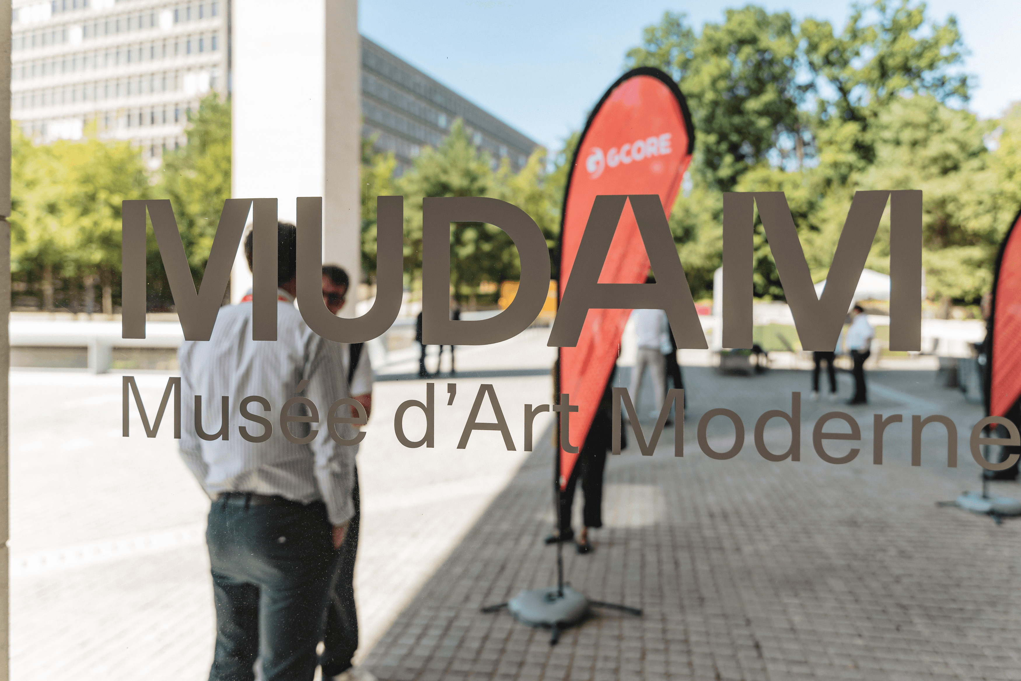 MUDAMI Musée d'Art Moderne text reflected on glass, with people and event banners inside.